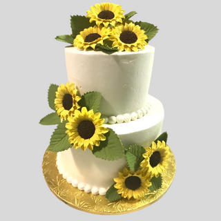 The Sunflower Tier is a two-tier white cake adorned with lifelike yellow sunflower and green leaf toppers, set on an embossed gold board—ideal as a bridal or engagement cake.