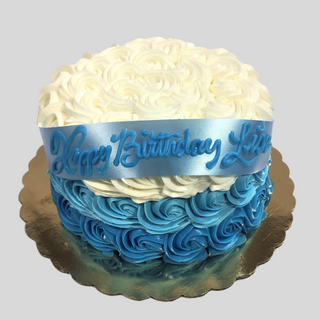 The Rosette Glam cake features a round ombre design with white and blue rosette frosting, a blue "Happy Birthday Lucas" ribbon topper, and a gold cake board—ideal for anyone seeking specialty birthday cakes.
