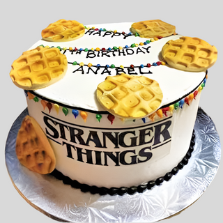 A round cake topped with icing Stranger Things Waffles, colorful Christmas lights, and "HAPPY BIRTHDAY ANAREL." The cake has the Stranger Things logo on the side—an ideal custom treat for kids!.