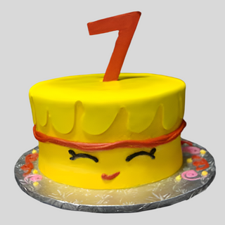 The Shopkins Number custom kids cake is a round yellow cake with a happy face, closed eyes, orange trim, a red number 7 topper, and sits on a silver board with colorful icing decorations.