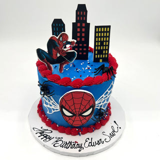 A blue Spider-Man City birthday cake features Spider-Man toppers, black spiders, web designs, city buildings, and "Happy Birthday Edwin Saul!" written on the white base.