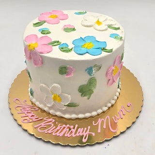 The Daisy Covered cake is a round cake with white frosting, featuring pink, blue, and white daisy designs on top. It sits on a gold cake board with "Happy Birthday Mum" written in pink icing.