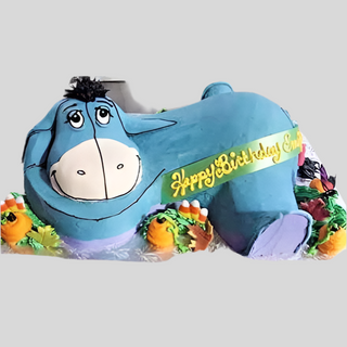 The Eore 3D cake is shaped like Eeyore from Winnie the Pooh, decorated with blue and purple icing, colorful carrots, and a yellow "Happy Birthday Emily" ribbon—perfect as a kids' cake for celebrations.