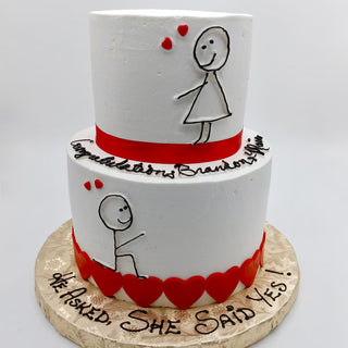 Figure Tier: A two-tier white engagement cake featuring red hearts and stick-figure proposal drawings. The base reads “He asked. She said YES!” with “Congratulations Brandon & Kylie” on the side.