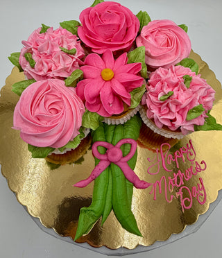 Moms Bouquet features cupcakes with pink and green frosting arranged like a flower bouquet on a gold board, with "Happy Mother’s Day" written in pink icing beside them.