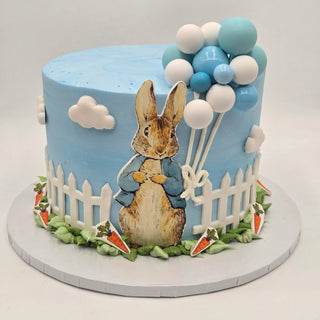 The Peter Rabbit Balloons cake is round and features a blue sky, white clouds, picket fence, a bunny holding blue and white balloons, with carrot decorations and green icing grass around the base.