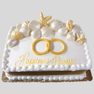 The Beach Rings cake is a rectangular white cake adorned with gold and white seashells, starfish, two gold engagement rings on top, and "Happiness Always" written in yellow—perfect for a bridal beach celebration.