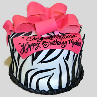 A custom round cake features zebra-striped black and white frosting, topped with the Zebra Bow and a pink plaque reading “Congratulations” and “Happy Birthday Mackie”—ideal for specialty birthday designs.