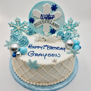 The Winter Onederland cake is white and blue, decorated with snowflakes, blue and silver balls, icing trees, and features “Winter ONEderland” and “Happy 1st Birthday Grayson” in blue icing.