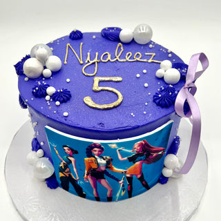 The K-Pop Glam cake is a purple birthday cake with white and purple icing, pearls, a purple ribbon, an edible image of three animated girls, and “Nyaleez 5” in gold on top.