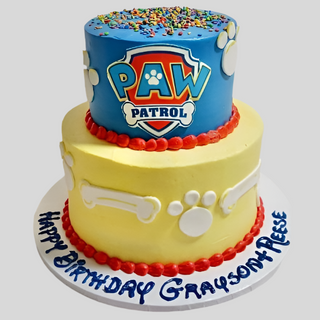 The Paw Patrol Tier is a two-tier custom kids cake with a blue top tier featuring paw prints, sprinkles, and the PAW Patrol logo, plus a yellow bottom tier with white bones. The base reads “Happy Birthday Grayson + Reese.”.