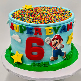 A round kids birthday cake decorated with Mario Sprinkles, yellow stars, clouds, and "SUPER EVAN," featuring Super Mario and a large red number 6. Blue and green icing add the finishing touch to this fun custom character cake.