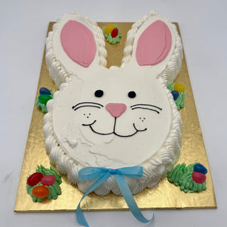 The Bunny Head cake features white frosting, pink ears, a smiling face, a blue ribbon bow, and colorful candy eggs on green grass-like frosting, all presented on a gold board.