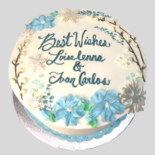 The Winter Bridal cake is a round white cake adorned with blue and white flowers, snowflakes, and branches, perfect for a winter engagement. It features “Best Wishes Luise Lenna & Ivan Carlos” written in blue icing.