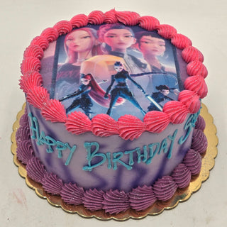 Round K-Pop Hunters cake with purple and pink frosting, featuring animated female characters on top. "Happy Birthday" is written in blue-green icing on the side. Cake sits on a gold-colored board.