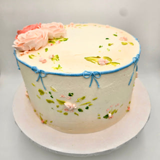 Simple Elegance is a round cake with light pastel icing, pink and green floral designs, blue piped bows on the sides, and pink buttercream flowers on top. Presented on a white cake board.