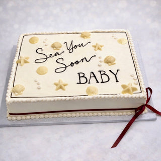 The Sea Baby is a rectangular white cake with gold seashells and starfish, “Sea You Soon Baby” in black script, pearl border, and accents of red ribbon and stripe.