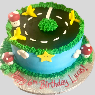 The Mario Mushrooms cake is a round treat topped with a grassy racetrack, yellow stars, red mushrooms with white spots, and white clouds. "Happy 6th Birthday Lucas!" is written in red icing.