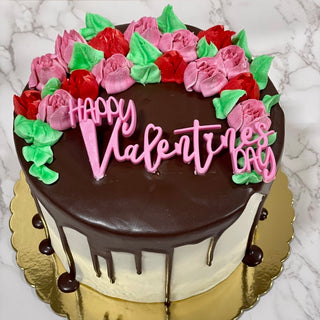 The Valentines Florals cake features white frosting with a chocolate drip, pink and red flower-shaped icing, green leaves, and a pink "Happy Valentines Day" topper on a gold cake board. Marble background.