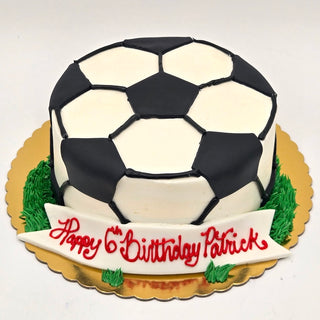 The Soccer Fondant cake features a soccer ball shape on a gold base, green icing "grass," and a white banner with “Happy 6th Birthday Patrick” in red—ideal for any soccer fan’s celebration!.