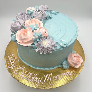The Colorful Celebration cake features light blue frosting, pink, lavender, and blue buttercream flowers on a gold board with “Happy Birthday Momma” in white icing.
