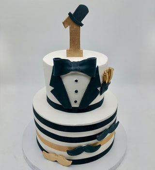 The Baby Bow-Tie cake is a two-tier creation with black and gold stripes, a gold mustache, bow tie, tuxedo jacket details, and topped with a gold number one and black top hat—perfect for a first birthday or anniversary celebration.