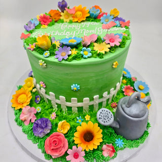 The Garden Beauty cake is green with colorful fondant flowers, a white picket fence, and a silver watering can. “Happy 77th Birthday Mom/Gma” is written on top. It’s displayed on a white surface.