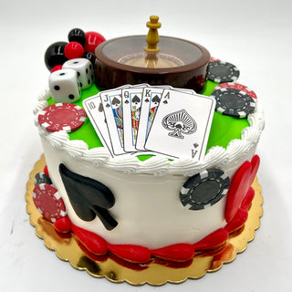 The Roulette Set is a round cake with white icing, red and black accents, decorated with poker chips, playing cards, dice, card suit symbols on the sides, and topped with a roulette wheel. Presented on a gold cake board.