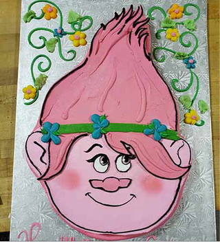 The Trolls Head kids' cake features tall pink hair, big ears, blue eyes, a wide smile with a green headband, and is decorated with colorful piped flowers and vines.