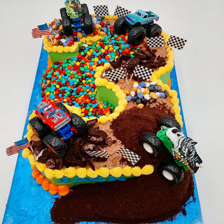 Monster Number cake features a colorful rectangular design with toy monster trucks, chocolate dirt, candy rocks, U.S. and checkered flags, recreating a racetrack on a vibrant blue base.