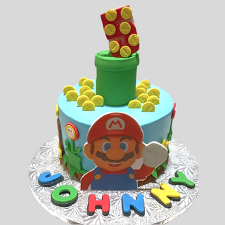 A round Mario Fondant cake decorated with Super Mario-themed designs: Mario fondant, gold coins, a green pipe, colorful blocks, and JOHNNY spelled in bright letters on the cake board—ideal as a kids cake!.
