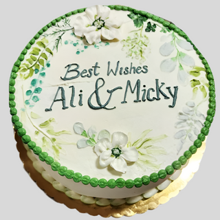 The Bridal Florals cake features a round shape, green border, white frosting, floral designs, and “Best Wishes Ali & Micky” written in green icing on top with painted leaves and white flower decorations.