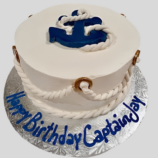 A round white cake topped with a Fondant Anchor and white rope, plus gold rings and more rope on the sides, with “Happy Birthday Captain Jay” in blue icing on the silver board.