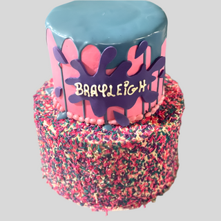 The Sprinkle Splat cake has two tiers: colorful sprinkles on the bottom tier and a blue and pink drip design above. The top features BRAYLEIGH in white on a purple splash—perfect for fun, character-themed kids' cakes!.
