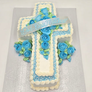The Religious Bouquet custom cross-shaped cake features white and blue icing, blue and green fondant roses for a Blue Floral accent, and a "God Bless Matthew" banner. The cake is displayed on a white surface.