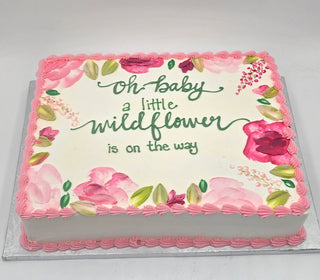 The Wildflower Baby cake is square, decorated with pink flowers and green leaves, and features the message “Oh baby a little Wildflower Baby is on the way” in green icing. Pink frosting borders add a charming finish.