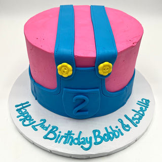 Ms. Rachel Overalls: A round pink cake with blue fondant overalls, yellow buttons, and a number 2 on the front. Presented on a white board with "Happy 2nd Birthday Bobbi & Isabella" in blue icing.