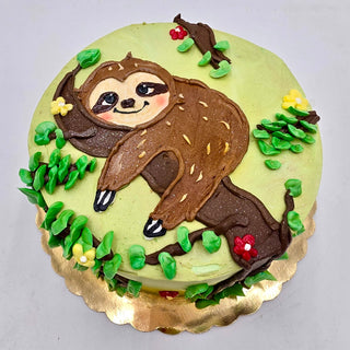 The "Hanging Sloth" cake features a frosted sloth lying on a branch, surrounded by green leaves with red and yellow flowers on a light green background, displayed on a gold board.