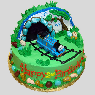 The Thomas Tunnel birthday cake for kids features a 3D train, tracks, and greenery. "Happy 2nd Birthday" is written in red and yellow icing, with candy rocks, a tree, and assorted playful characters completing the design.