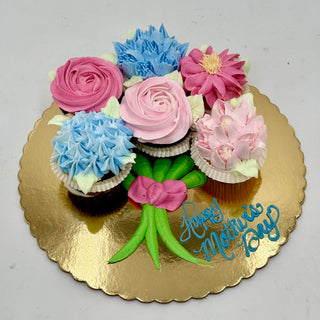 Bouquet Beauty features six cupcakes arranged as a bouquet with vibrant flower frosting, green icing stems, and “Happy Mother’s Day” written in blue icing on a gold board.