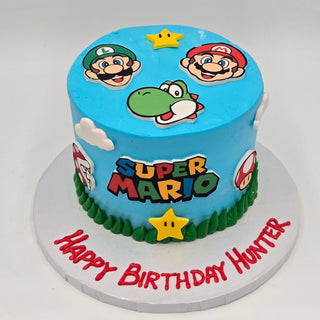 A blue kids birthday cake with a Super Mario theme, featuring the Mario Logo, yellow stars, mushrooms, clouds, and "Happy Birthday Hunter" on the cake board.