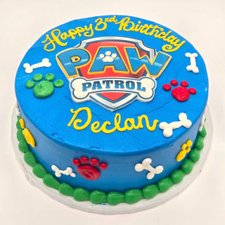 A blue Paw Patrol Scan kids birthday cake, decorated with colorful paw prints, bones, and "Happy 3rd Birthday Declan" in yellow icing, featuring the PAW Patrol logo at the center.
