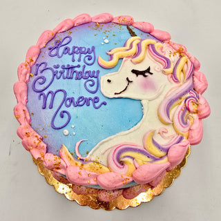A round cake decorated with Unicorn Frosting, featuring a pastel unicorn and pink border. “Happy Birthday Maeve” is written in purple on a blue background with white and gold accents.
