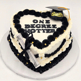 Heart Degree cake features black and white frosting, gold pearl accents, a fondant graduation cap, diploma decor, and is topped with “ONE DEGREE HOTTER” in bold black letters.