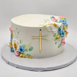 Artistic Religion: A round white cake adorned with vibrant buttercream flowers and leaves, highlighted by a gold cross on the side. Presented on a white and silver base against a simple background.