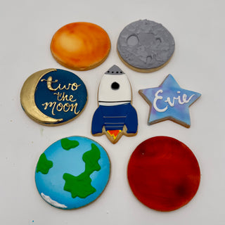 Space Cookies: A set of seven decorated cookies featuring planets, a rocket, a star with "Evie," and a crescent moon with "to the moon" in gold icing, all arranged on a white background.