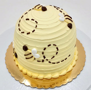 Bee Smash is a round, dome-shaped yellow cake with smooth frosting, chocolate lines, and small bee designs on top. It features piped icing accents and a bee-themed look, presented on a gold cake board.