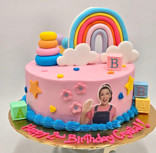 A pink birthday cake decorated with Ms. Rachel Toys—rainbow, clouds, stacking rings, blocks, stars, pink flowers—and an edible image of a smiling girl waving. Text reads “Happy 2nd Birthday Crystal!”.