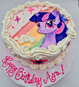 A round custom Pony Rainbow cake with a purple cartoon pony, white icing swirls, gold sprinkles, pink stars, and "Happy Birthday Ava!" written in pink frosting on the cake board.