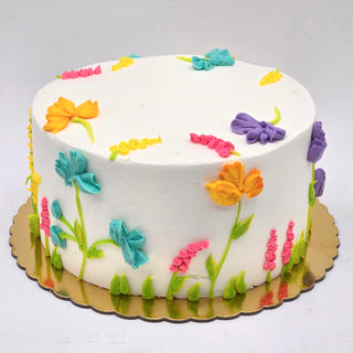 The Wildflower Assorted cake is a round white custom birthday cake, decorated with colorful buttercream wildflowers and green stems, presented on a gold board—perfect for specialty birthday designs.
